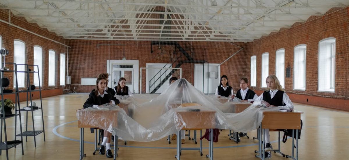 A group of students in uniform sit around desks covered in plastic in a large gymnasium.