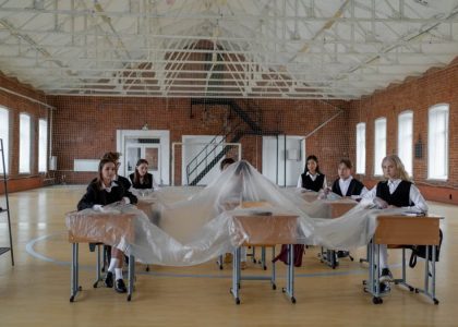 A group of students in uniform sit around desks covered in plastic in a large gymnasium.